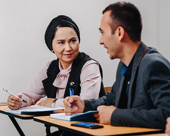 Female Webster student with braided hair talking during a class discussion with a fellow student behind her.