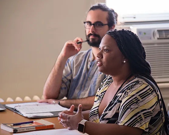 Two students sitting in a classroom; one is speaking, the other is listening closely